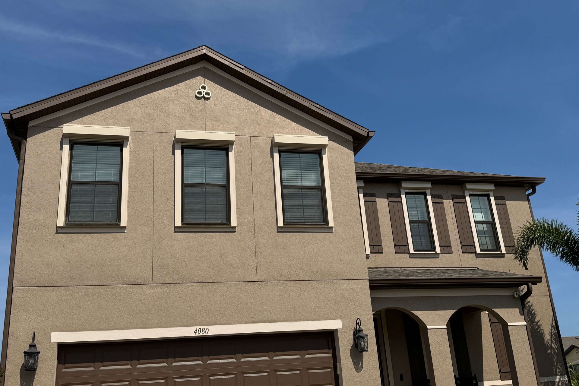 A tan, two-story house with a brown garage door, arched porch entry, and three windows across the upper level.