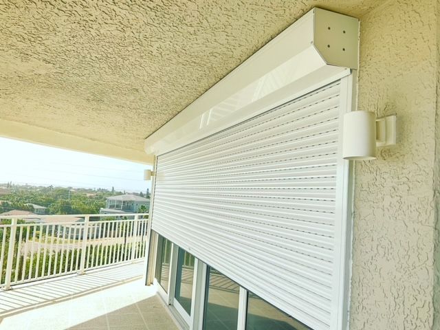 A white exterior rolling shutter covers a glass sliding door on a balcony overlooking a residential area.
