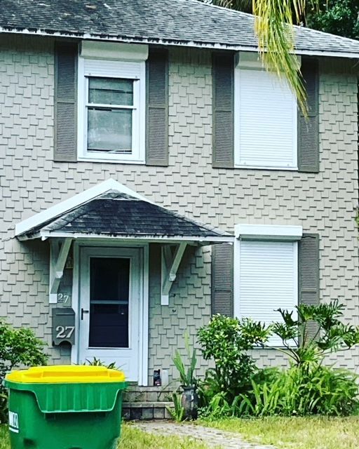 A two-story grey brick house with white-framed windows, some with security shutters, and a green trash bin in the yard.