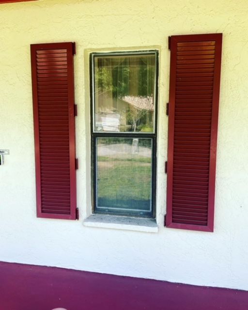 A window with dark red louvered shutters on either side, set against an off-white stucco exterior wall.