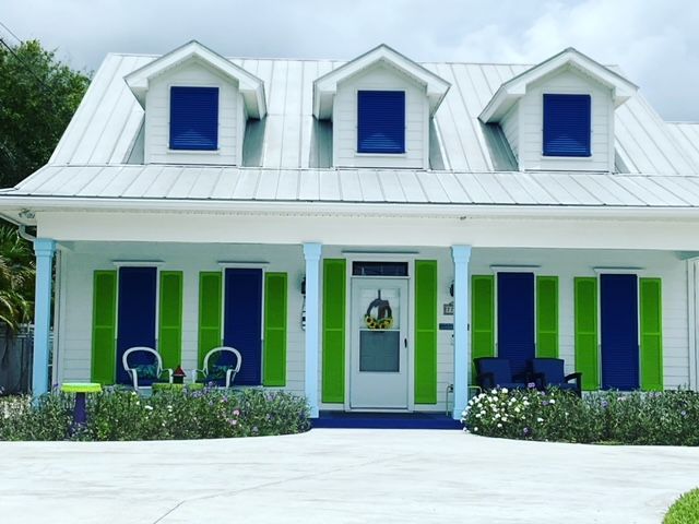 A one-story white coastal house with a metal roof, three dormers, blue storm shutters, and bright green accents.