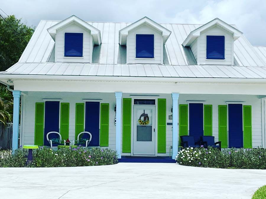 A white one-story cottage with a metal roof, three dormers, and bright blue and green window shutters on a sunny day.