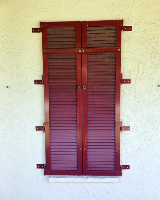 A pair of dark red, louvered shutters closed over a window on a textured white stucco wall.