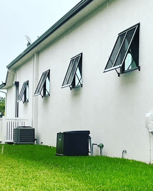 Exterior view of a white wall with four black awning windows open at an angle above two HVAC units on a green lawn.