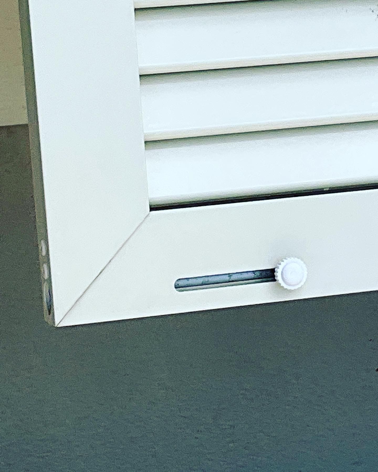 A white metal shutter corner with a small, adjustable circular knob set within a horizontal sliding track.