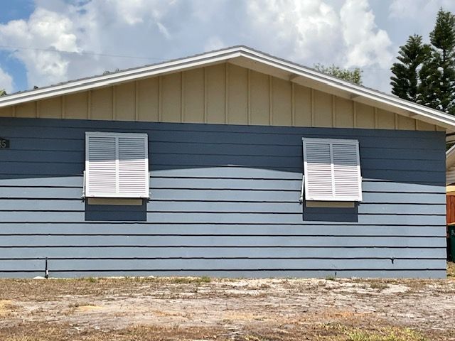 A single-story house with blue horizontal siding, tan vertical gable siding, and two white shutters on a sunny day.
