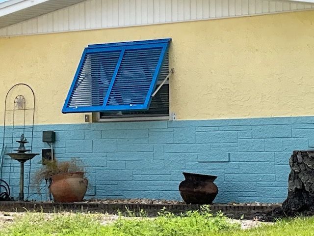 A blue, louvered storm shutter is propped open on a yellow and blue house wall above a terracotta pot and birdbath.