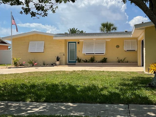 A single-story, yellow house with white hurricane shutters, a green-trimmed front door, and an American flag on the lawn.