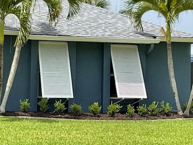 Two white bahama shutters pushed open on a blue house exterior, framed by palm trees and a landscaped garden bed.