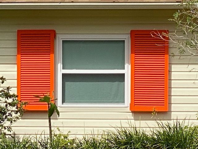 A white window with bright orange shutters mounted on a beige, horizontal-sided house wall.