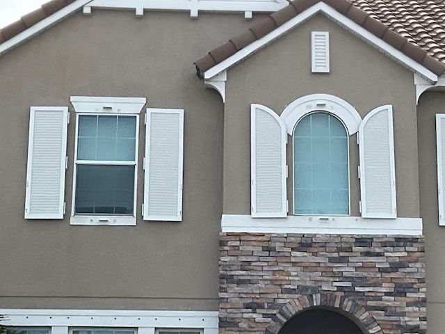 The exterior of a tan stucco house features a stone veneer, white window trim, and arched window shutters.