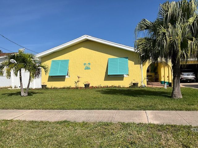 A bright yellow, single-story house with turquoise window shutters, a front lawn, and palm trees under a blue sky.