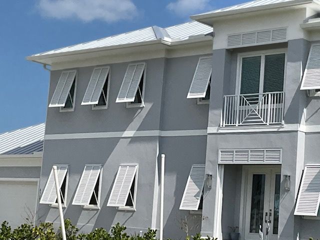 A two-story grey stucco house featuring white hurricane shutters, a metal roof, and a balcony under a bright blue sky.