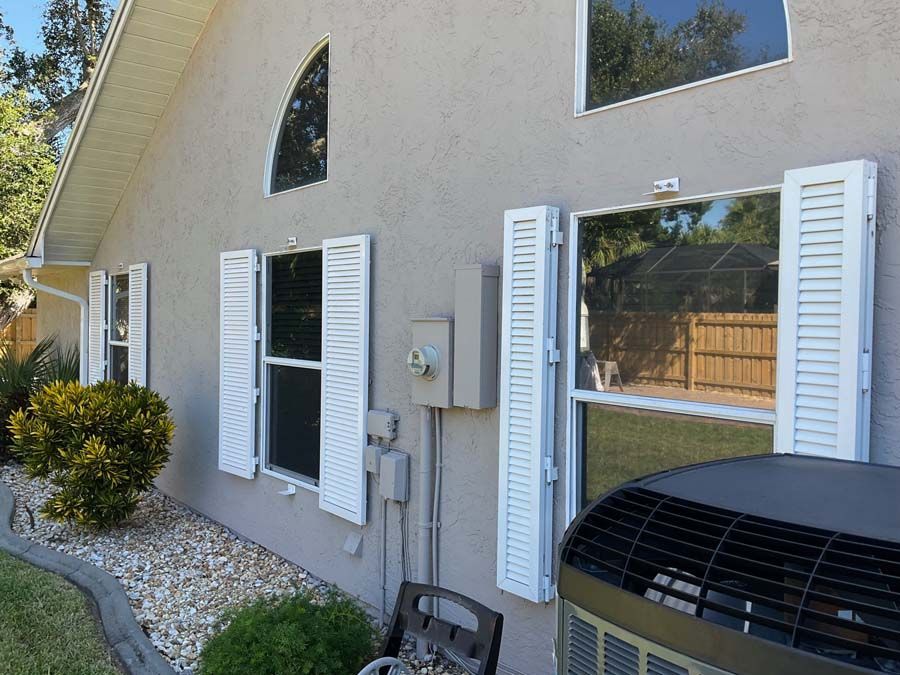 A beige house exterior with white-shuttered windows, an arched window, electrical boxes, and an outdoor AC unit.