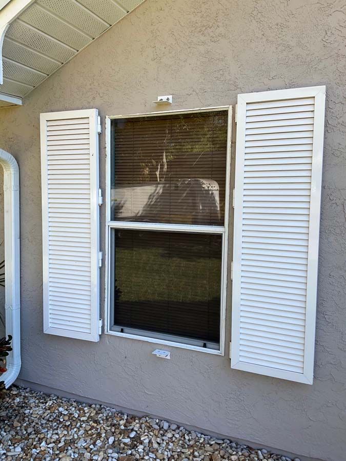A beige exterior wall with a window flanked by two white, louvered shutters and decorative hardware below the frame.