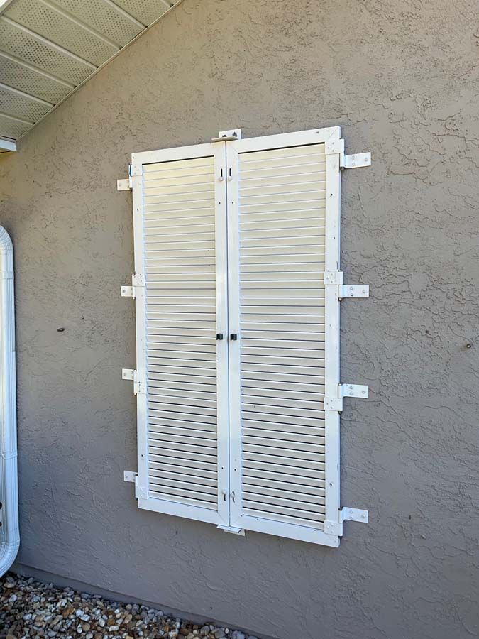 White louvered storm shutters closed over a window on a textured stucco exterior wall.