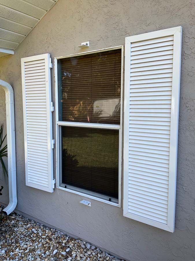 A rectangular window with white louvered shutters on a beige stucco wall, viewed from an exterior angle.