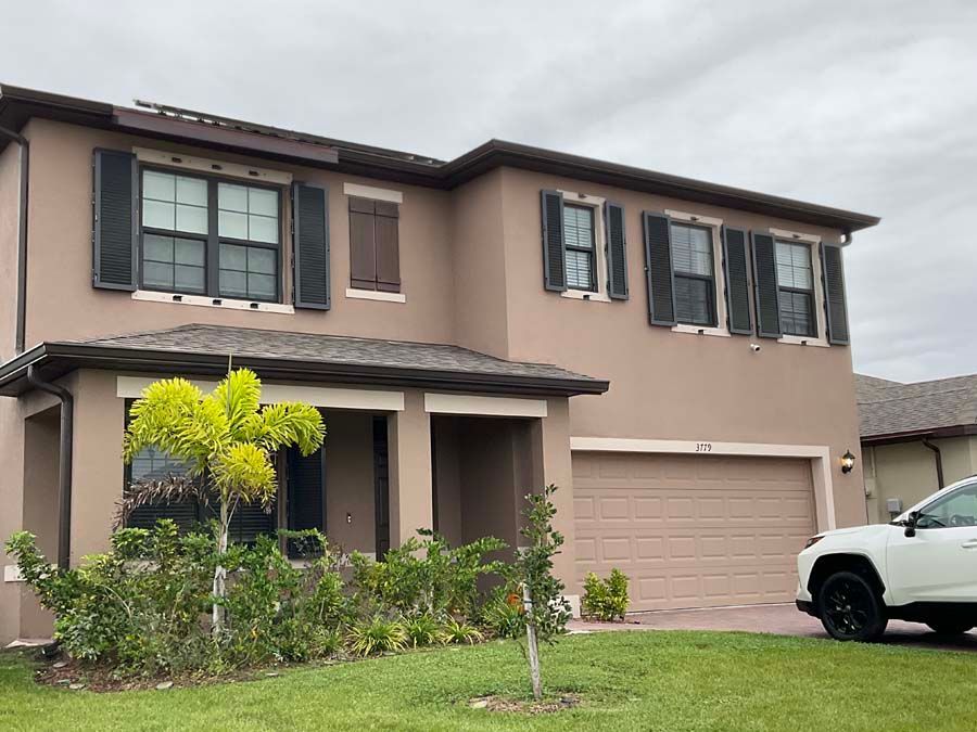 A two-story, tan suburban house with dark shutters, a front porch, a two-car garage, and a palm tree in the front yard.