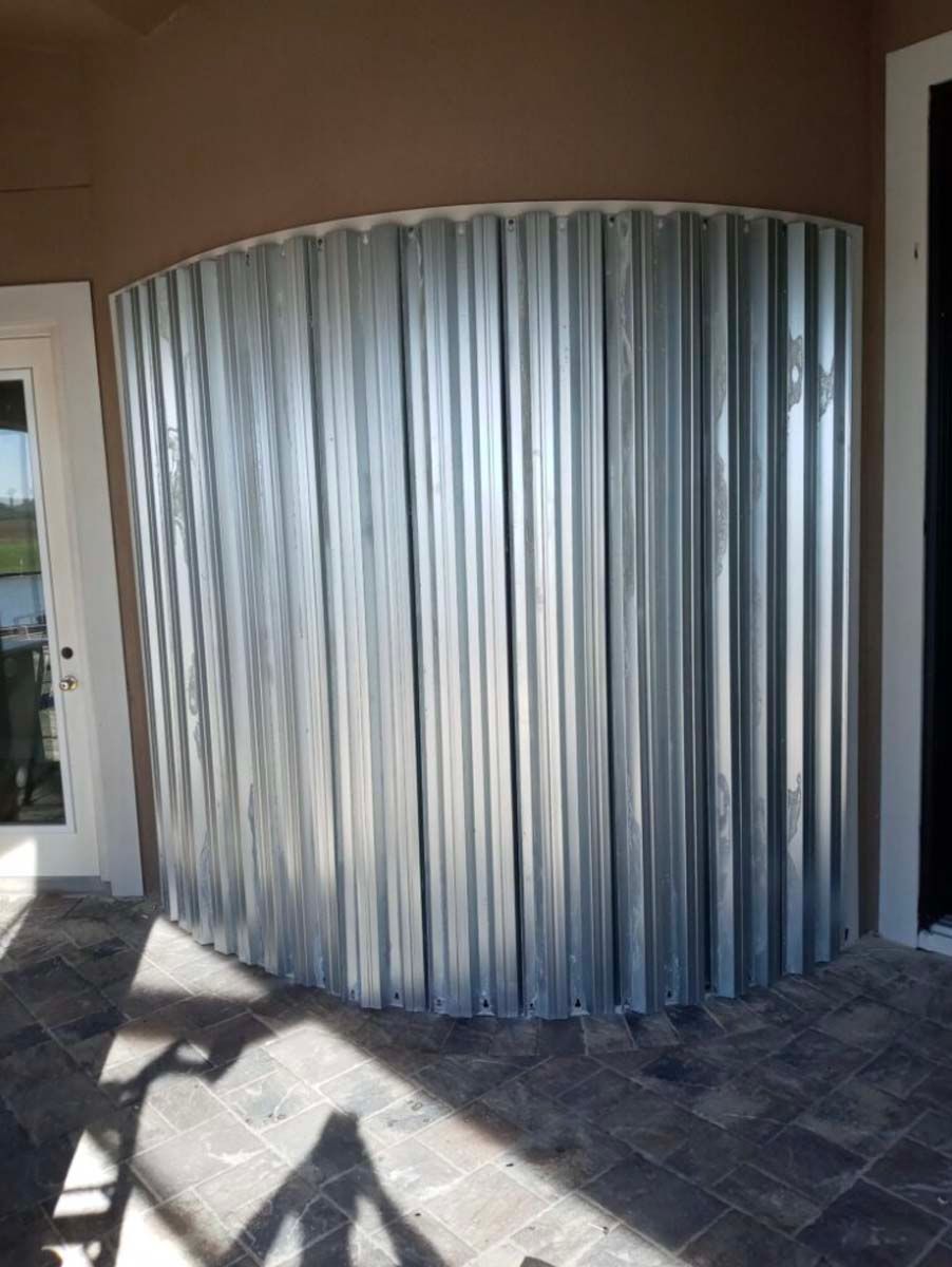 Silver corrugated metal accordion storm shutters installed on a curved outdoor patio wall.