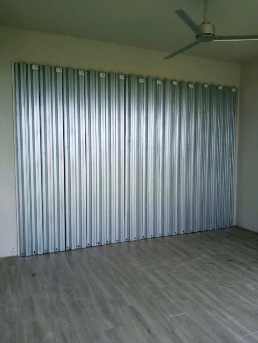 A silver vertical accordion-style partition door covering a wall in a room with light-colored flooring and a ceiling fan.