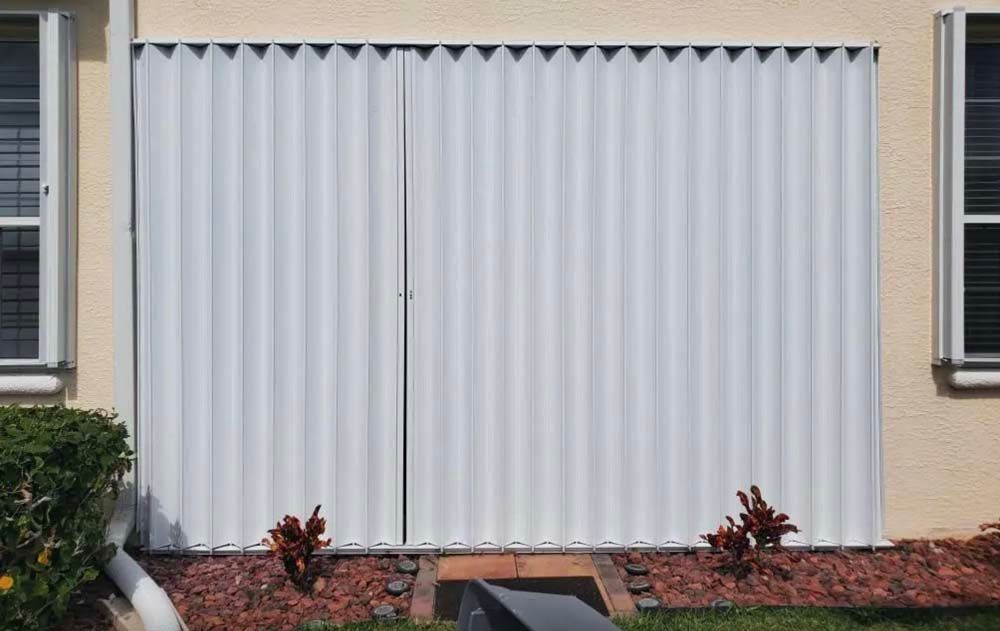 White accordion hurricane shutters fully closed over a doorway on a light-colored stucco house exterior.