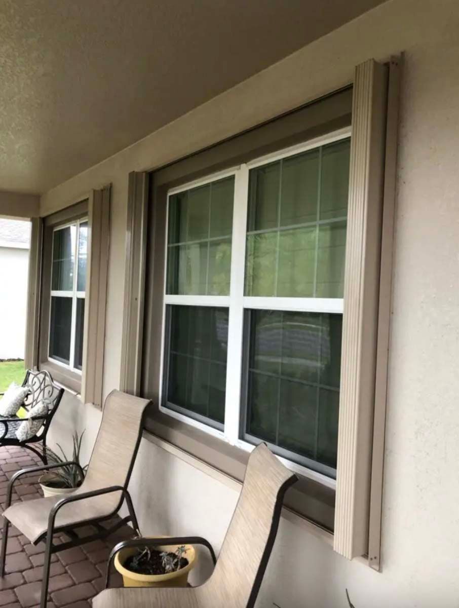 Two brown outdoor patio chairs sit on a brick porch beneath windows with beige shutters on a light-colored stucco wall.