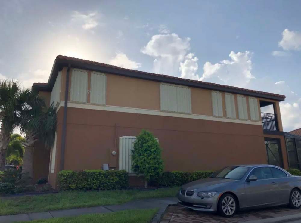 A two-story tan and brown condo exterior with hurricane shutters on the windows, parked car, and landscaping.