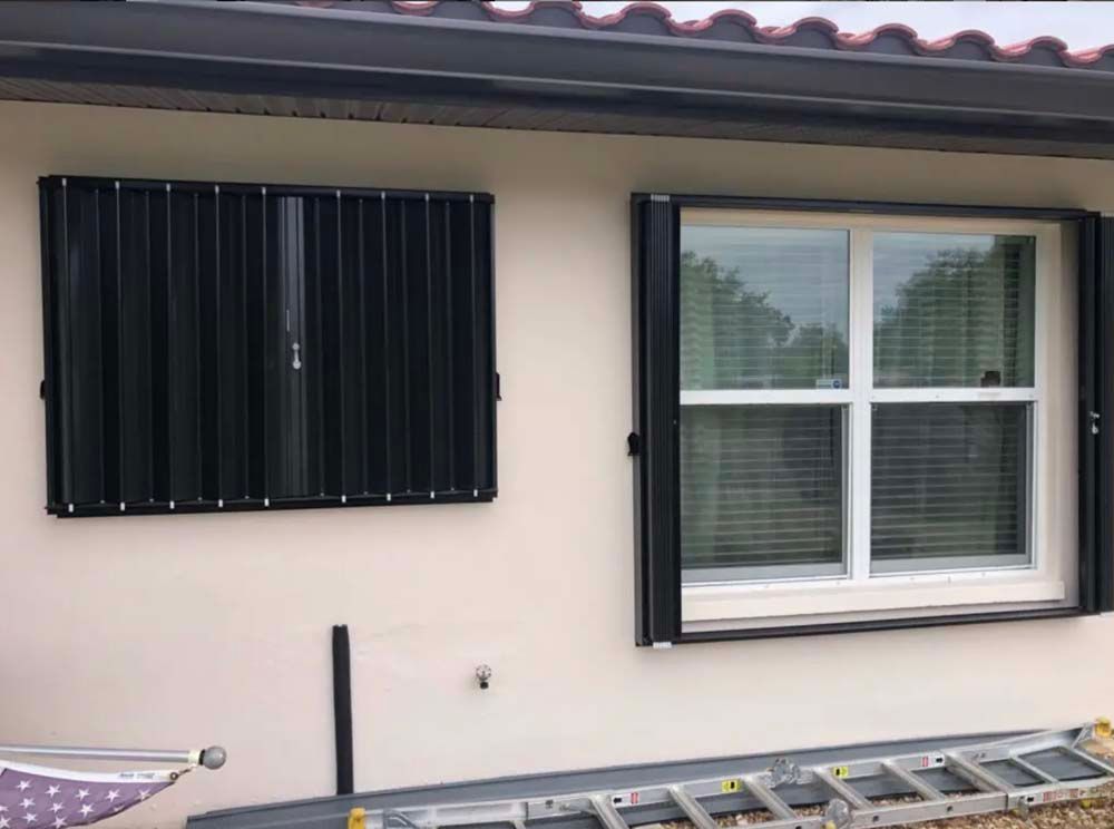 A house wall featuring a closed black hurricane shutter on the left and an open window with its shutter frame on the right.