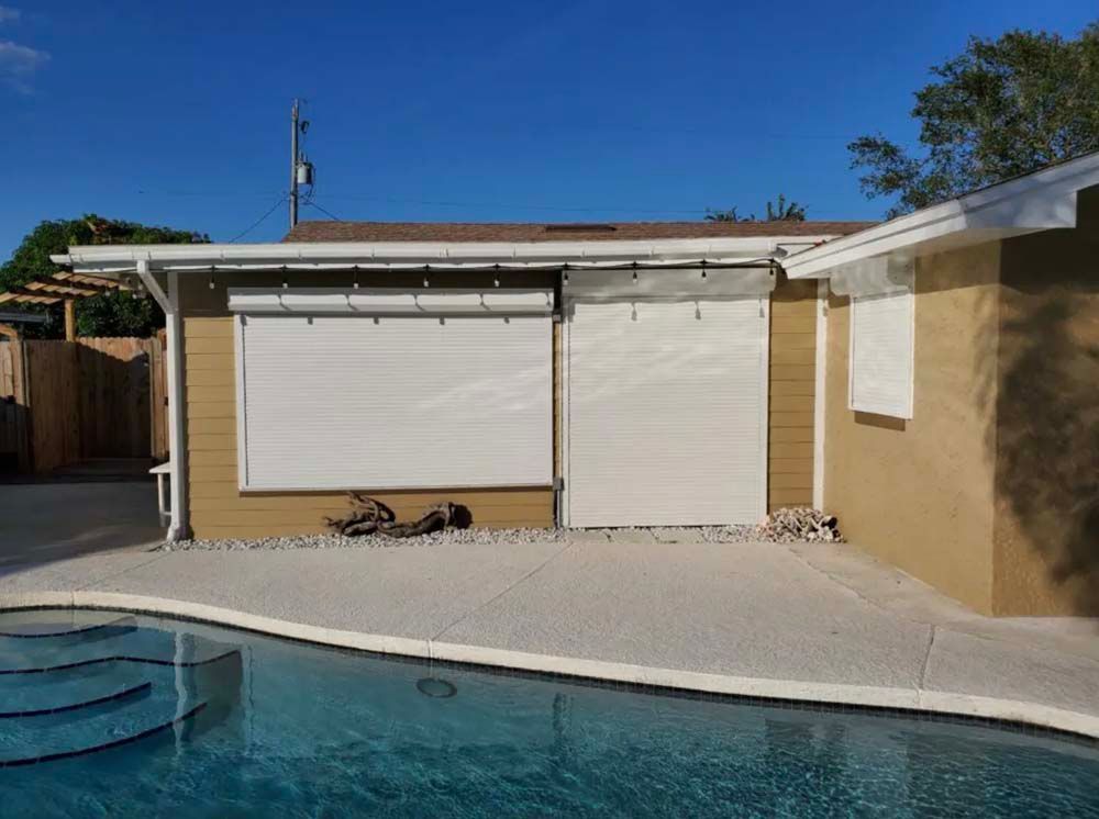 A tan house exterior with beige outdoor shades pulled down over windows next to a swimming pool under a blue sky.