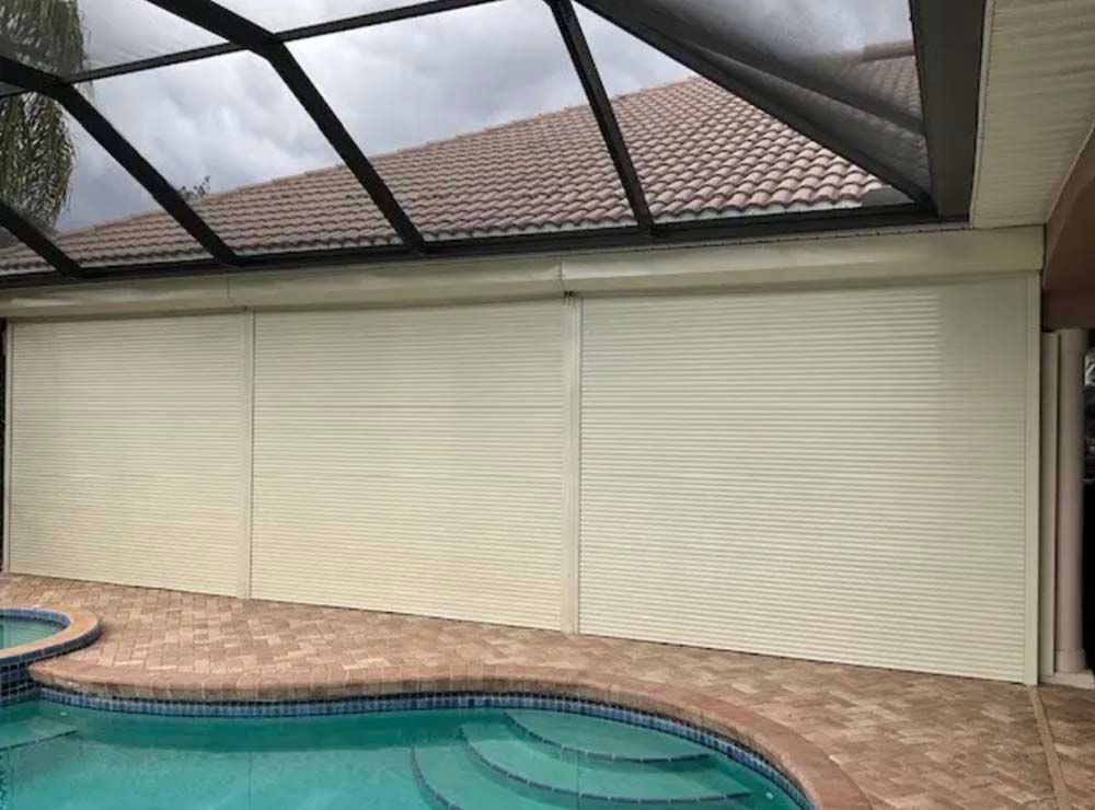 Off-white roll-down storm shutters covering a pool enclosure wall under a screen roof and tiled roof.