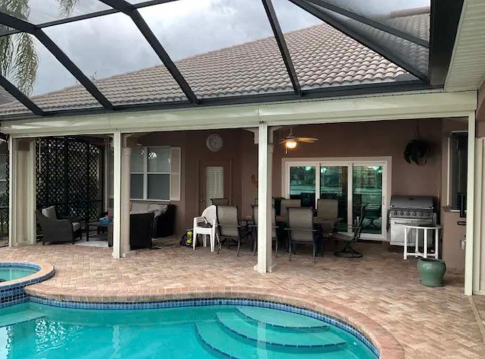 A covered patio featuring outdoor dining furniture and a grill, adjacent to a backyard swimming pool.
