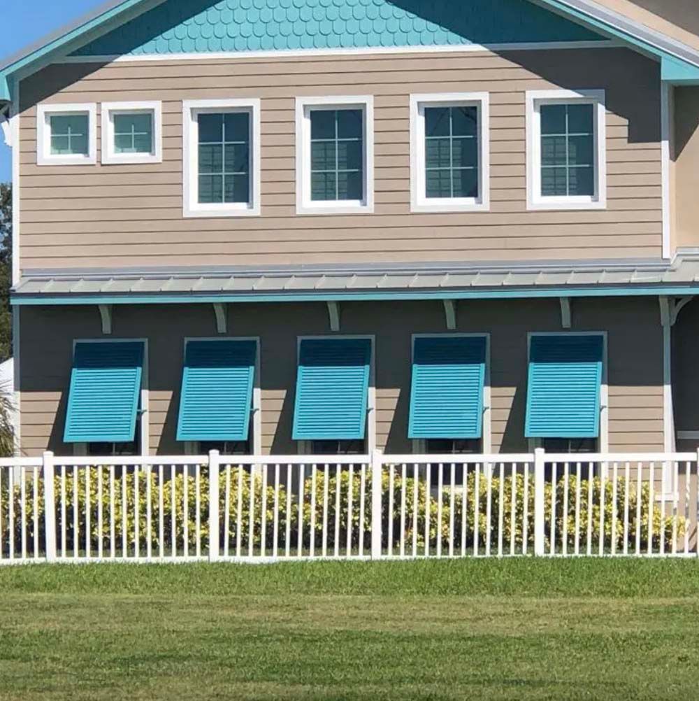 A two-story tan house with a turquoise roof, blue window awnings, and a white picket fence in front of a green lawn.