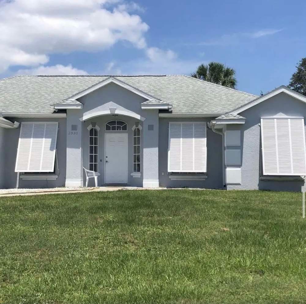A single-story gray house with a white door, light gray roof, and white shuttered windows under a bright blue sky.