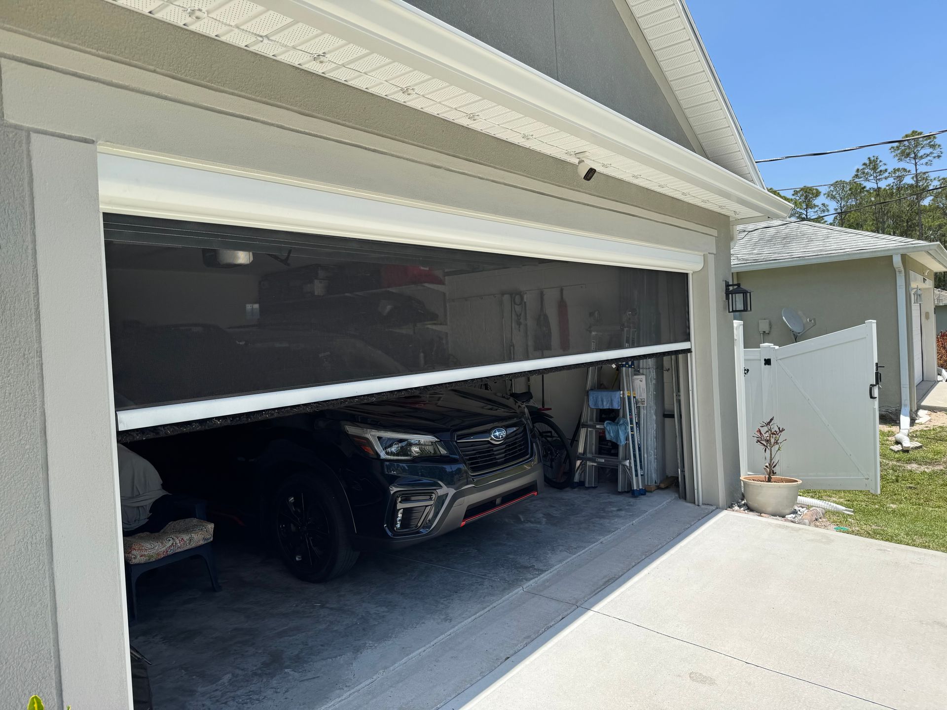 A black garage door screen partially covers an opening with a dark vehicle parked inside a garage on a sunny day.