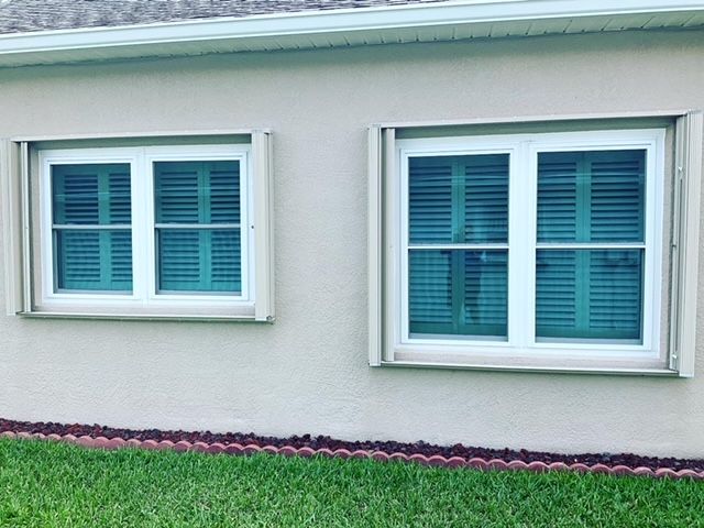 Two white windows with teal shutters installed on a light-colored stucco exterior wall above a grass lawn.
