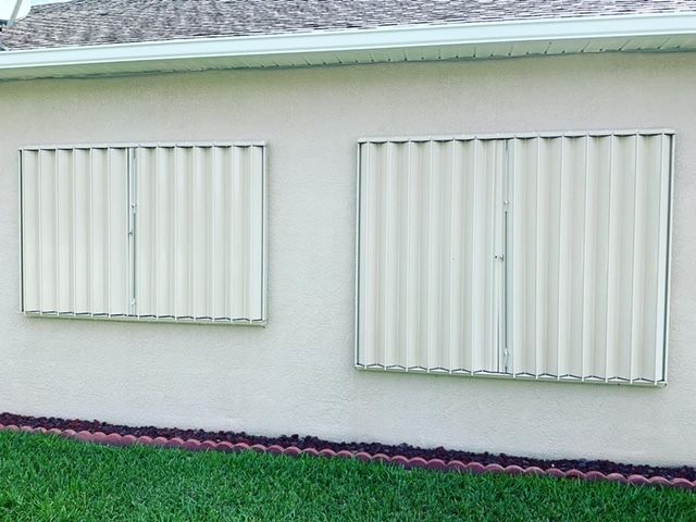 Two beige accordion-style hurricane shutters installed over windows on a light-colored exterior wall.