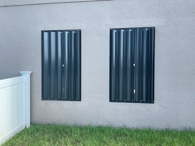 Two dark metal accordion hurricane shutters are closed over windows on a light-colored stucco exterior wall.