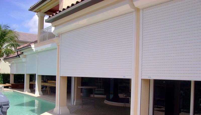 White rolling storm shutters are partially lowered over a residential patio and swimming pool area.