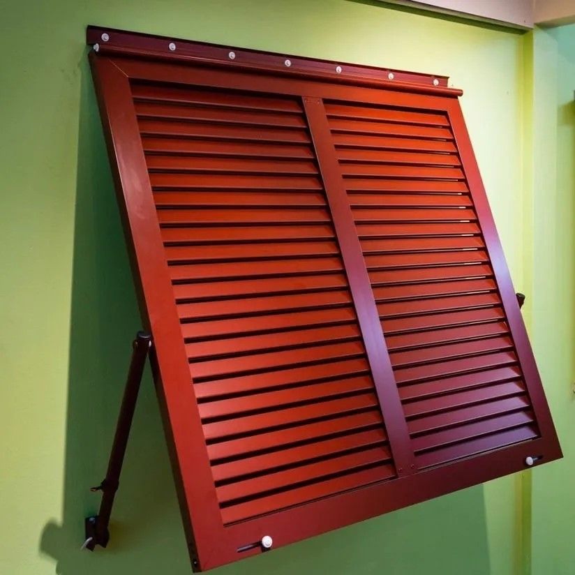A red, louvered window shutter mounted on hinges, propped open at an angle against a light green wall.