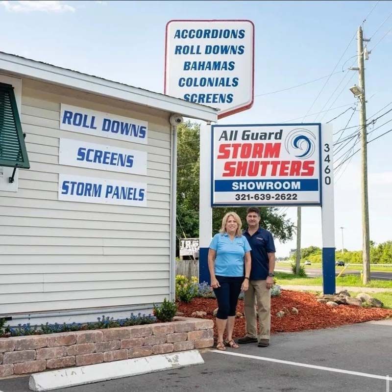 Two people stand outside All Guard Storm Shutters, a business with informative signage on the building and a tall sign.
