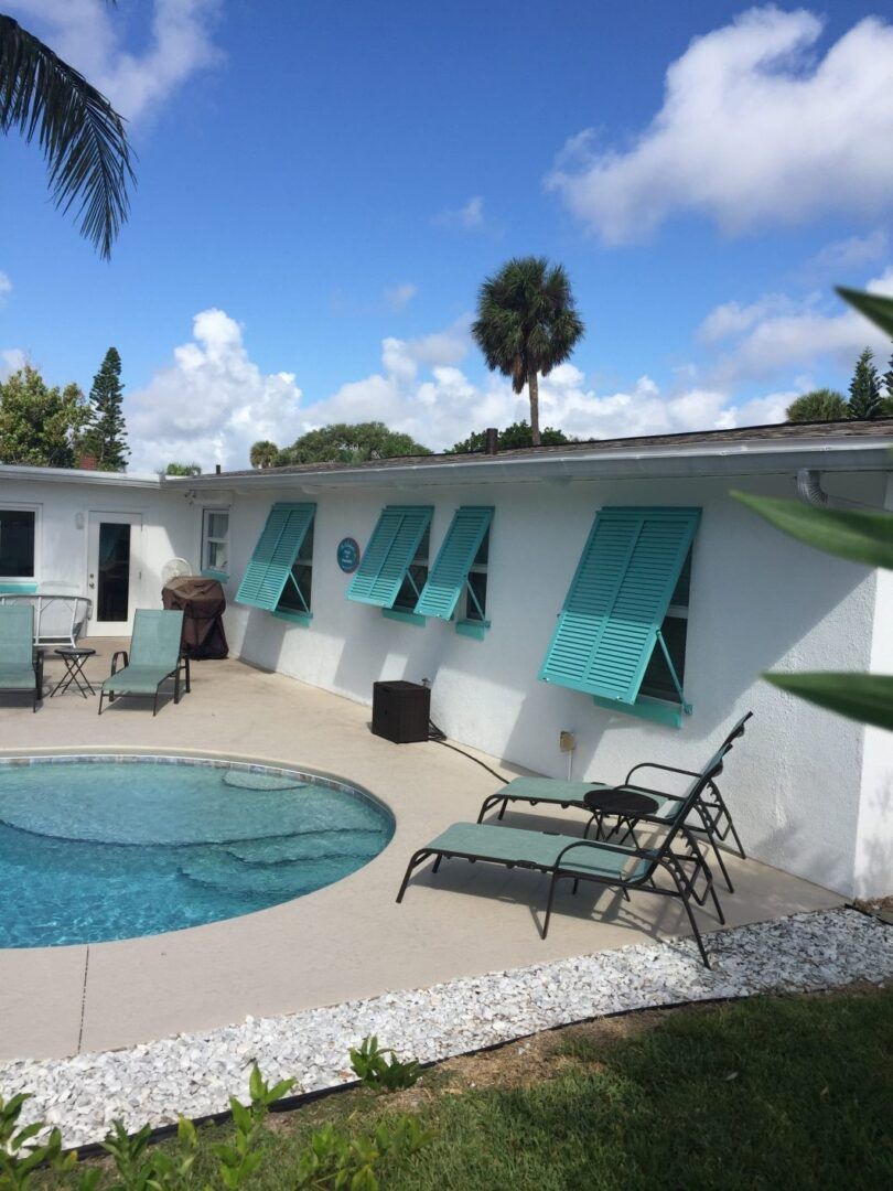 A backyard pool with lounge chairs sits beside a white house featuring distinctive turquoise shutters under a sunny sky.