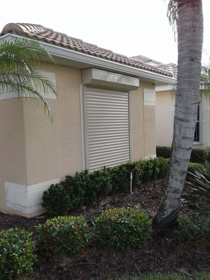 A beige exterior wall of a house features a closed, horizontal-slatted metal hurricane shutter over a window.