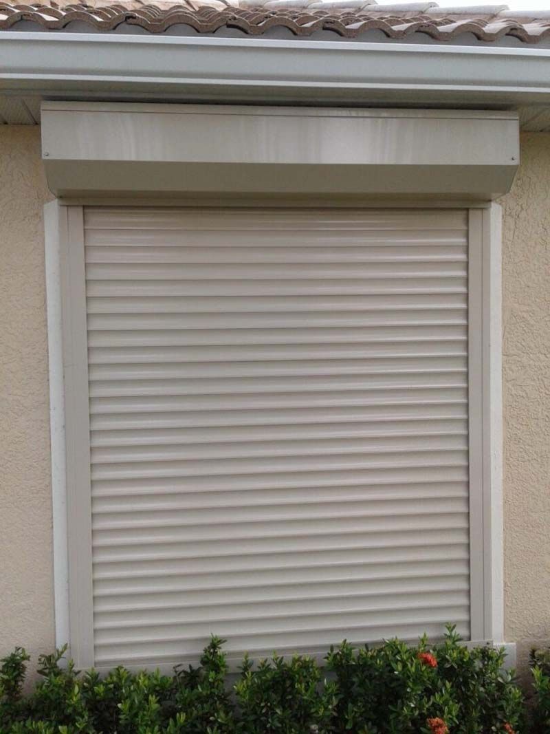 A beige exterior window with a closed, horizontal-slat security rolling shutter, positioned above a small hedge.