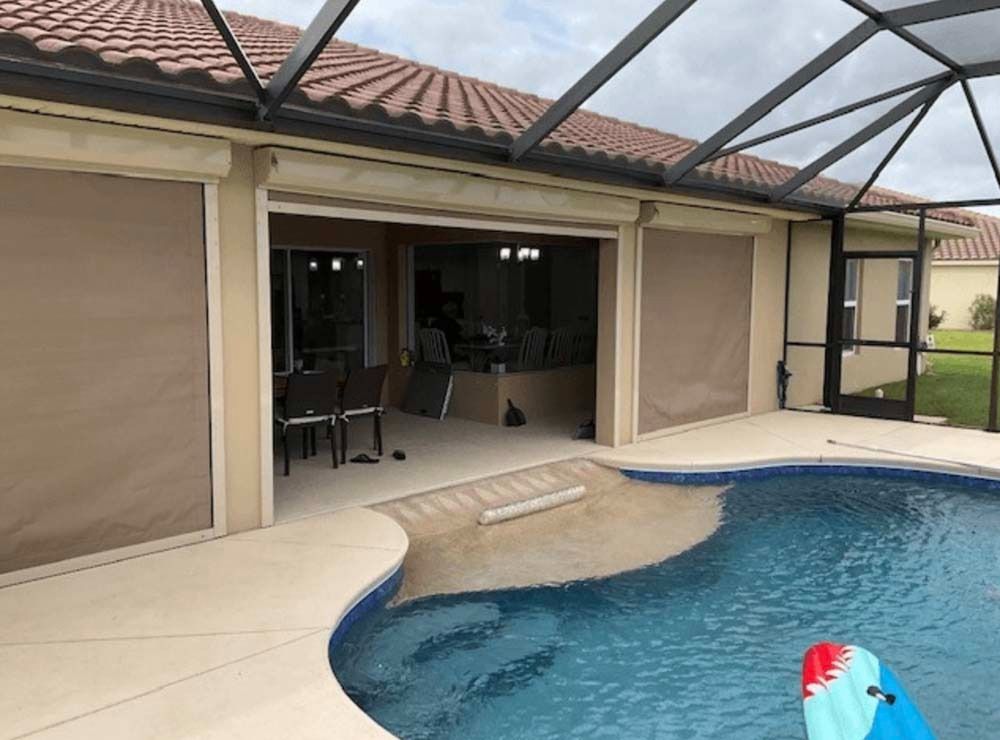 A patio with tan retractable shades, pool, and screen enclosure under a cloudy sky.