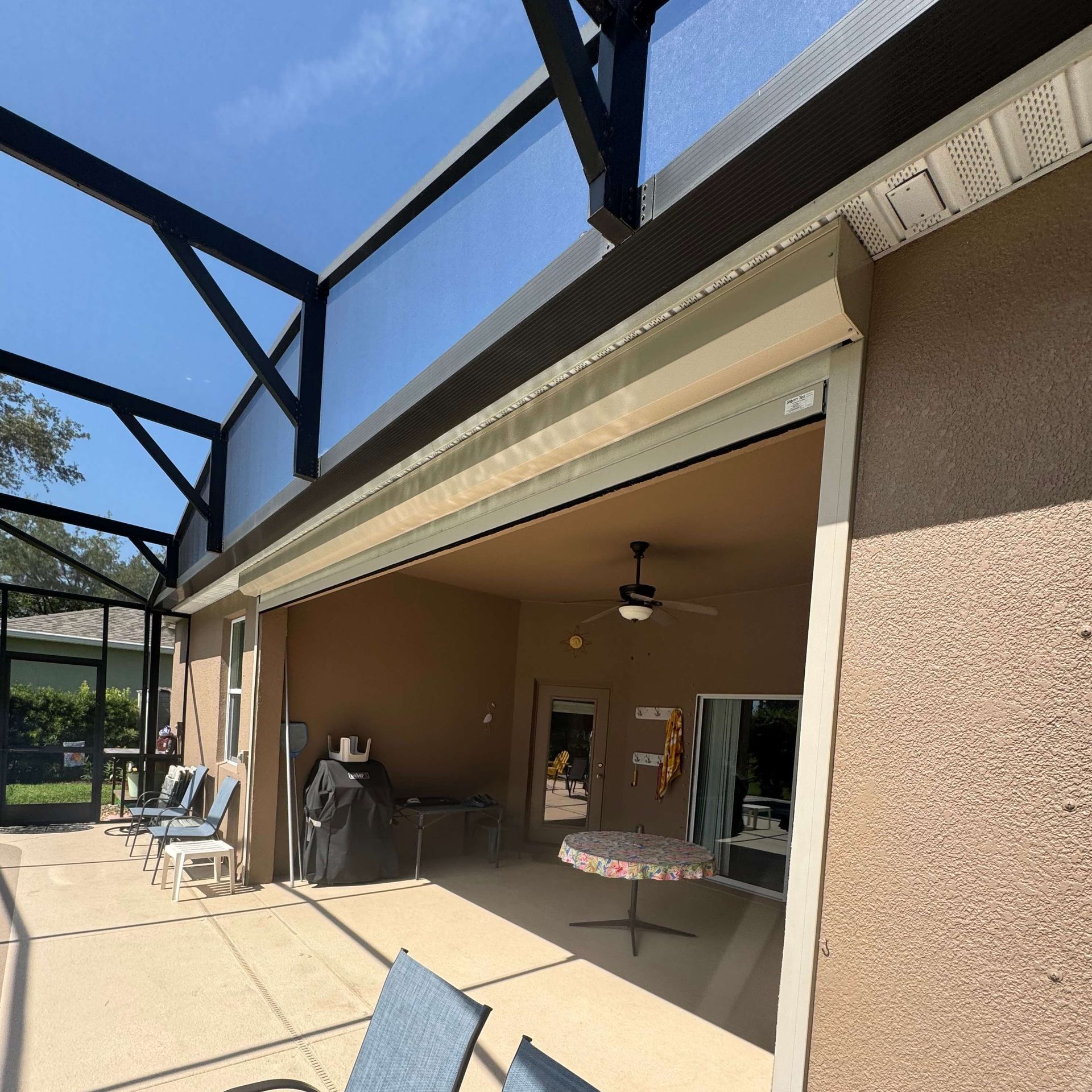 A covered patio with a partially lowered beige motorized screen, outdoor furniture, and a ceiling fan.