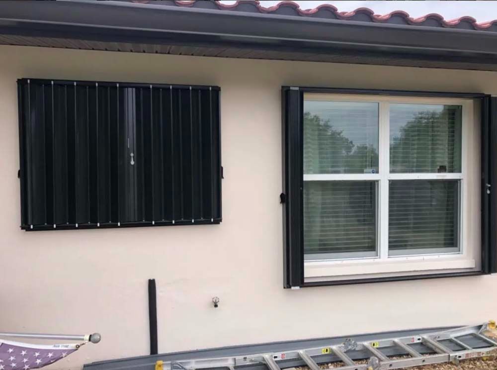 A beige exterior wall with a black accordion hurricane shutter closed on the left and open on the right beside a window.