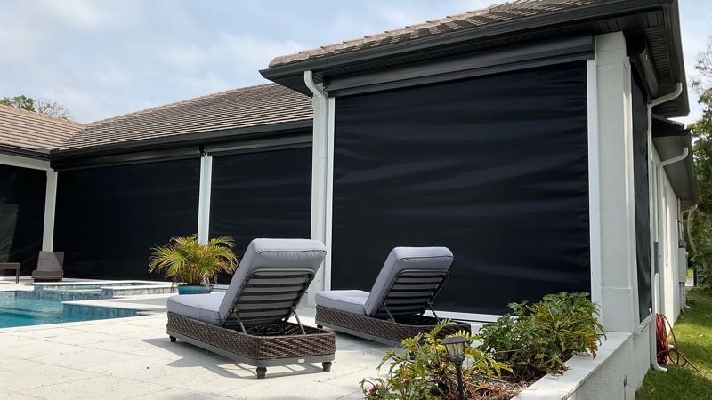 Two lounge chairs on a light-colored pool deck under a roofed patio area with installed black privacy screens.