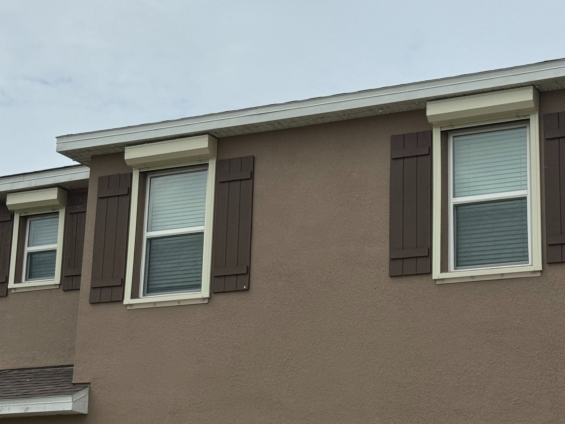 Upper level of a beige stucco house exterior with three windows featuring brown shutters and white storm shutters above.