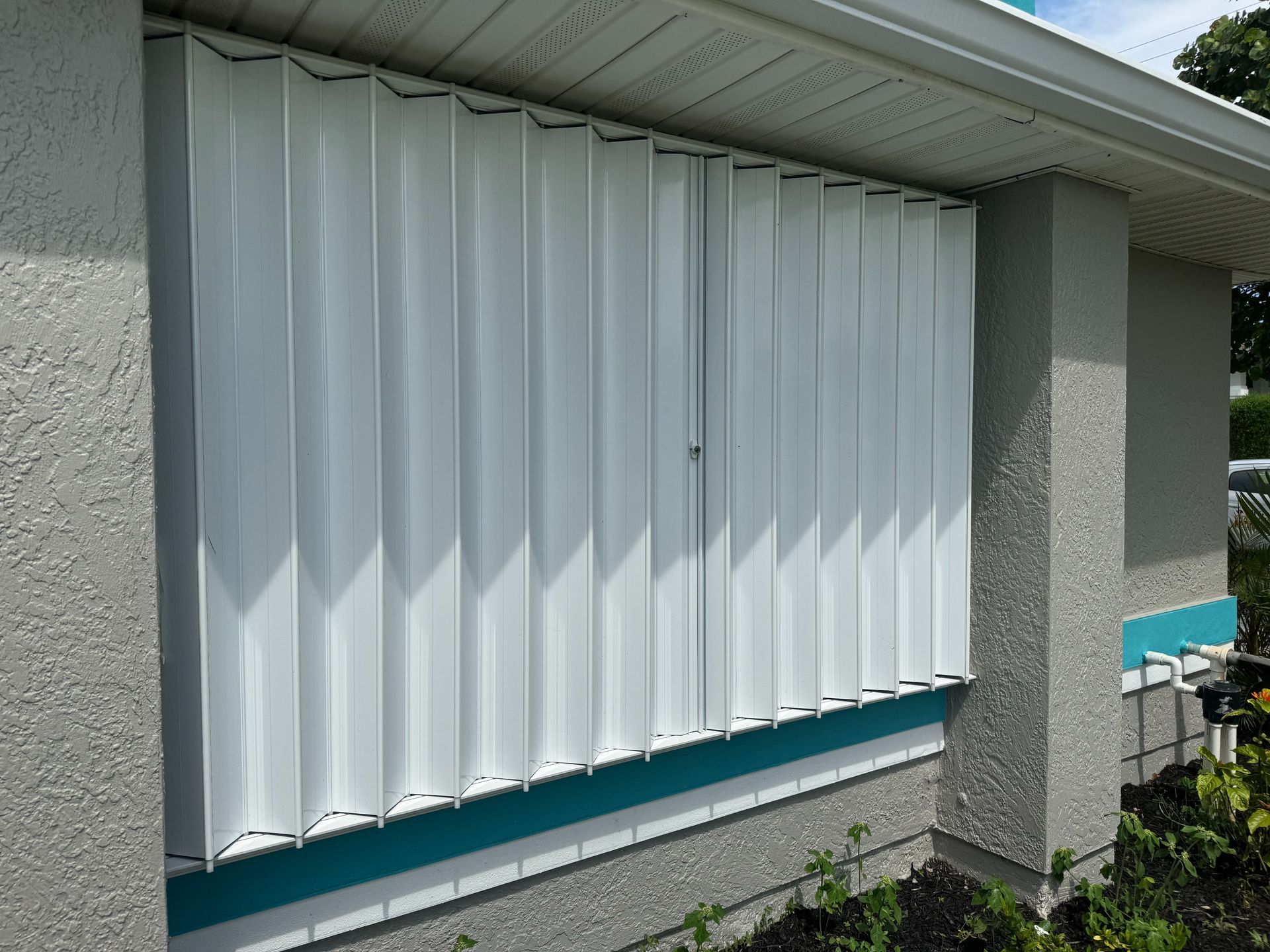 White accordion-style storm shutters closed over a window on a textured, light-colored house exterior.