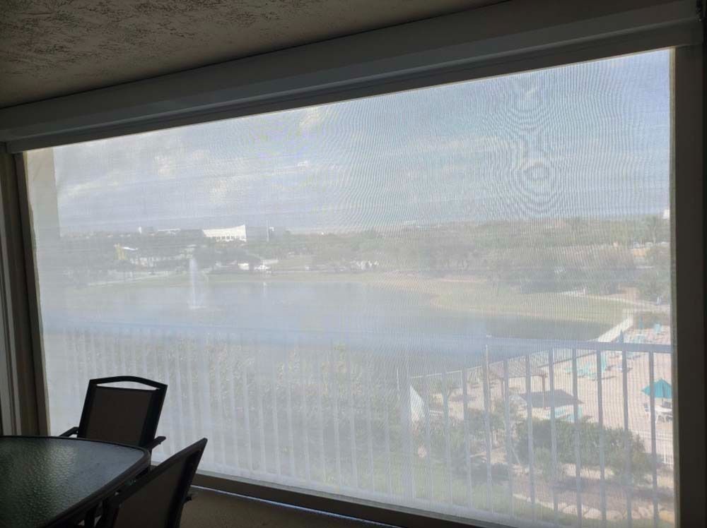 A window with a light solar shade pulled down, looking out over a lake, green landscape, and balcony railing.